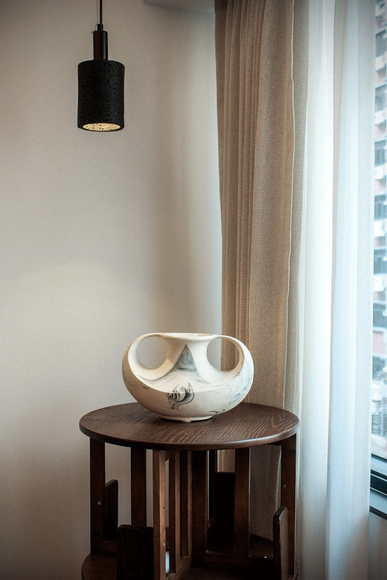 Living room corner with ceramics on a round timber table under a black pendant and beige curtains